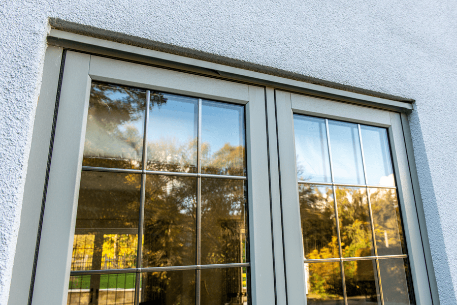 Close-up of a green flush sash window with reflective glass panes, featuring a grid pattern, installed in a textured white exterior wall, capturing the reflection of nearby trees and sky.