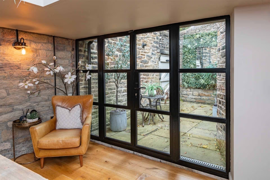 A cozy indoor seating area featuring Decorio steel-look aluminium doors with multiple panes, leading out to a stone-walled courtyard. The room has a natural stone wall, light wood flooring, a tan leather armchair, and a decorative branch with white flowers.