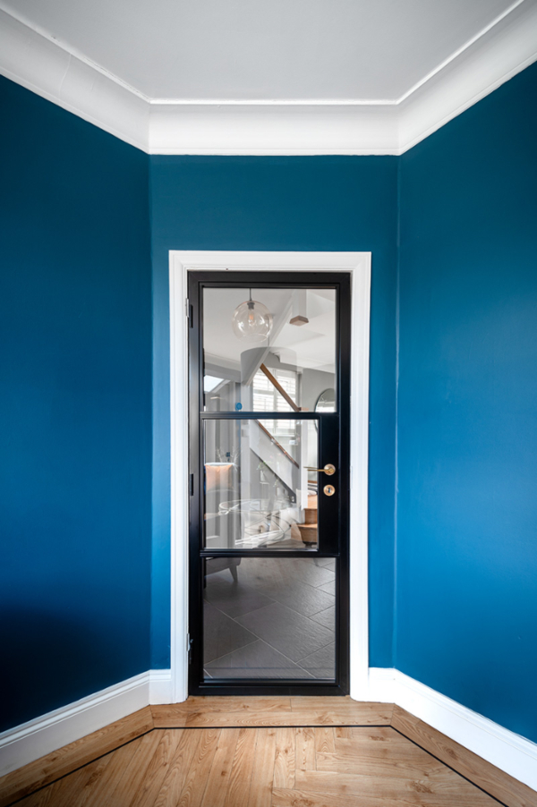 A single, black Decorio steel-look aluminium internal door with multiple clear glass panes, set in a corner of a room painted a vibrant, deep blue. The door has a prominent handle and provides a clear view into the adjacent hallway with a staircase and dark flooring, contrasting with the light wood floor in the foreground.
