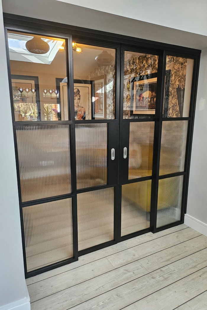 A set of black Decorio steel-look aluminium sliding internal doors with multiple panes, featuring a combination of clear glass in the upper sections and textured, fluted glass in the lower sections for privacy. The doors open into an interior room with patterned wallpaper and artwork, and are set against a light wood-effect floor.