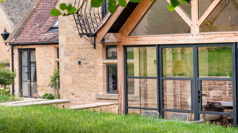 A bright dining area featuring Decorio steel-look aluminium doors and an apex sidelight, set within a vaulted ceiling with exposed timber beams. The room has light wood flooring, a long wooden dining table with fur-covered chairs, and views of a garden patio.
